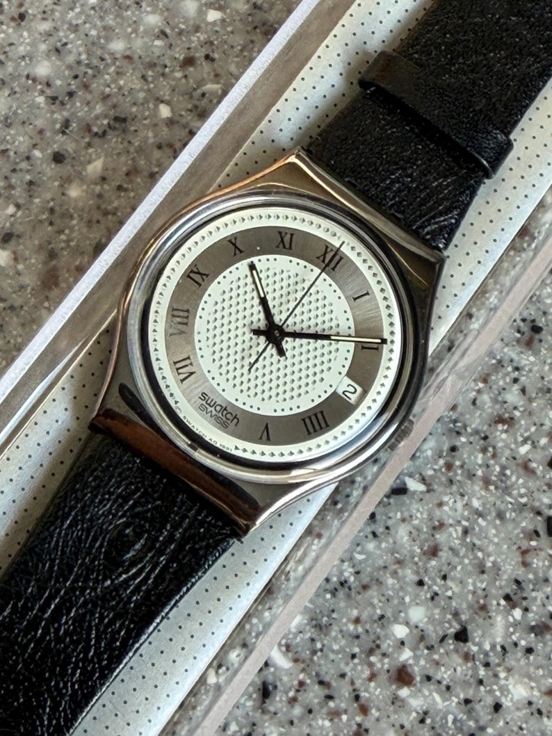 K&ouml;nnte beinhalten: Eine Swatch-Uhr mit silberfarbenem Geh&auml;use und schwarzem Lederarmband. Das Zifferblatt der Uhr ist wei&szlig; mit schwarzen r&ouml;mischen Ziffern und Zeigern. Die Uhr befindet sich in der Originalverpackung.