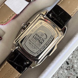 Puede incluir: Un reloj Invicta Lupah plateado con una correa de cuero negro. La esfera del reloj es plateada con marcas negras y el texto "COS INVICTA LUPAH THE LAST WORD OY SOLO MOLEMINES Spacial Edition TRITNITE&trade; FLAME-FURY CRYSTAL WATER RESISTANT MODEL NO. 5168" est&aacute; impreso en la parte posterior.
