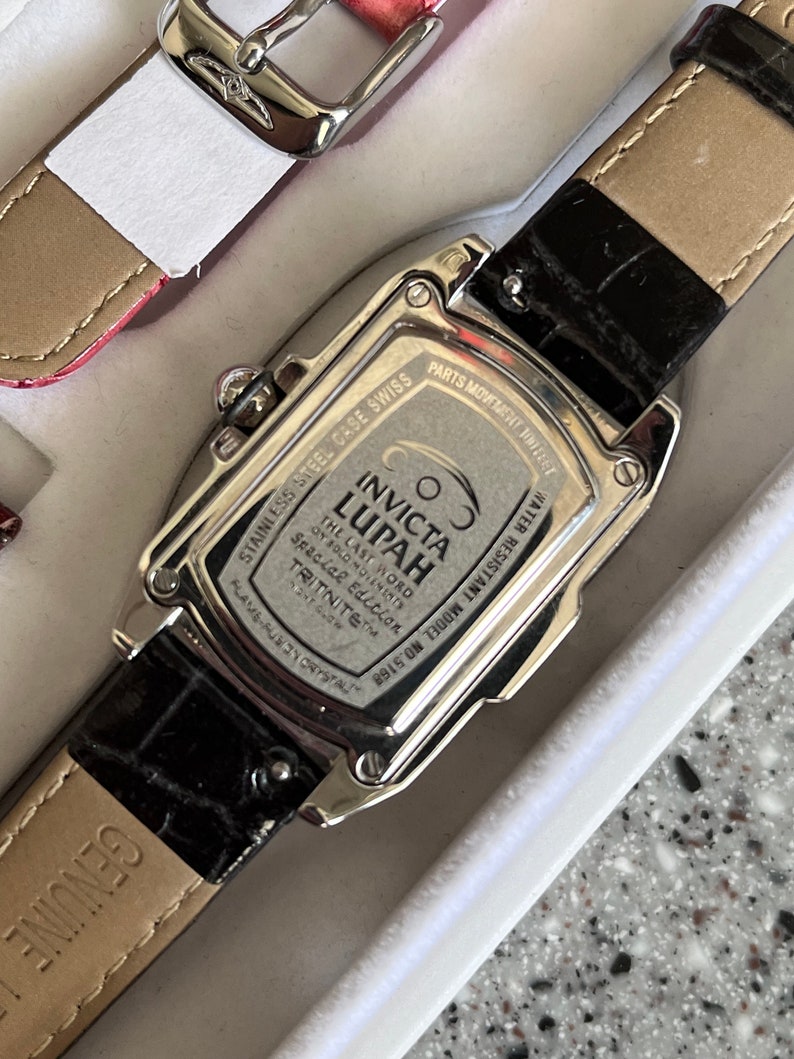 Puede incluir: Un reloj Invicta Lupah plateado con una correa de cuero negro. La esfera del reloj es plateada con marcas negras y el texto "COS INVICTA LUPAH THE LAST WORD OY SOLO MOLEMINES Spacial Edition TRITNITE&trade; FLAME-FURY CRYSTAL WATER RESISTANT MODEL NO. 5168" est&aacute; impreso en la parte posterior.
