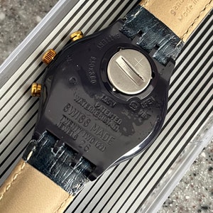 May include: A black Swatch watch with a gold-tone face and a black leather band. The watch has a water-resistant feature and is made in Switzerland. The back of the watch has the text "Swatch Genuine Leather Made in Switzerland" and "Waterresistant Patented Swiss Made Twenty Two (22) Jewels 26".