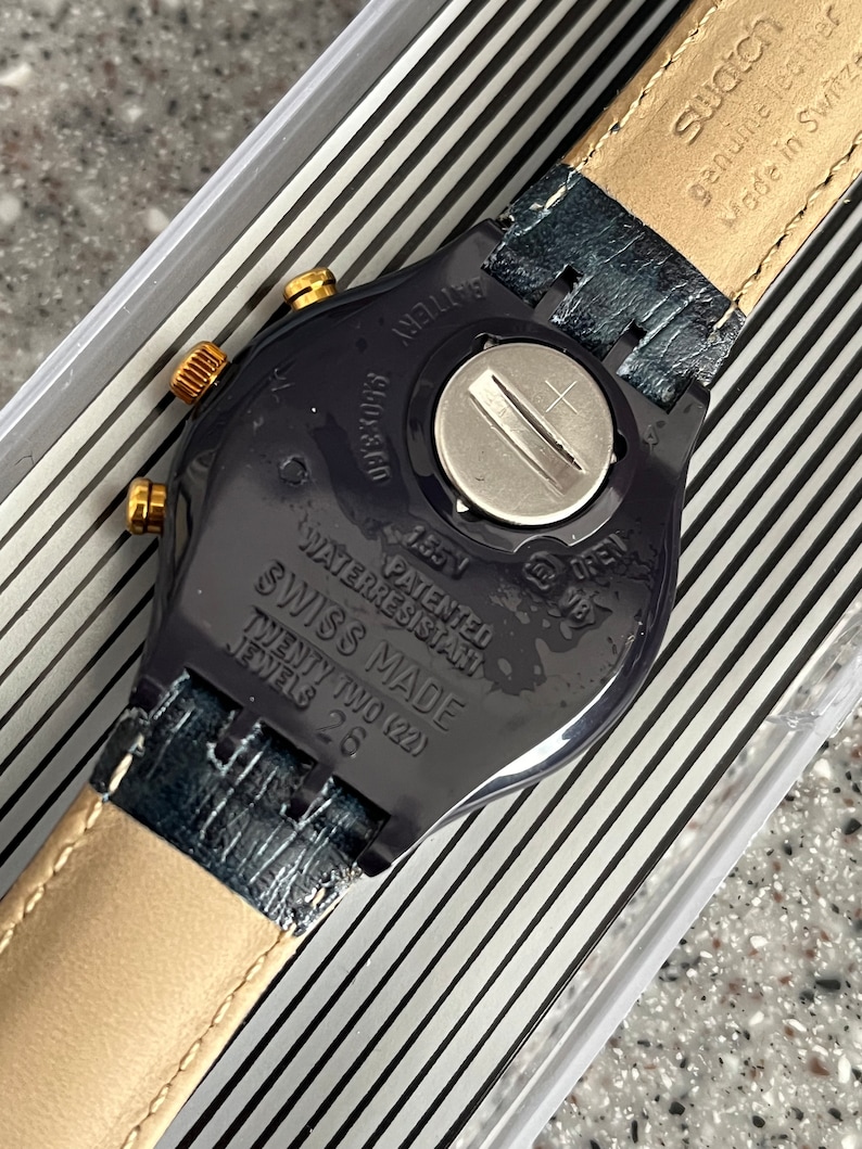 May include: A black Swatch watch with a gold-tone face and a black leather band. The watch has a water-resistant feature and is made in Switzerland. The back of the watch has the text "Swatch Genuine Leather Made in Switzerland" and "Waterresistant Patented Swiss Made Twenty Two (22) Jewels 26".