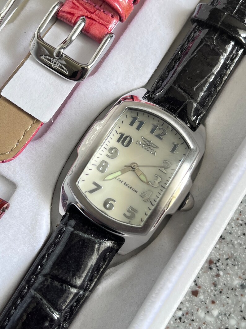 Puede incluir: Un reloj plateado con una correa de cuero negro y una esfera blanca. La esfera del reloj tiene marcadores de hora en n&uacute;meros romanos negros y las palabras "Flat Edition" impresas en ella.