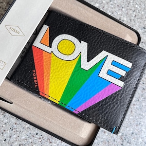 Puede incluir: Cartera de cuero negro con la palabra "LOVE" en letras blancas y un diseño de arcoíris. La cartera está en una caja Fossil. La cartera es de estilo plegable.