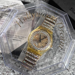 May include: A gold-tone Swatch watch with a clear face and a crystal-encrusted band. The watch is in a clear plastic box with the words "Hollywood Dream" printed on the side.