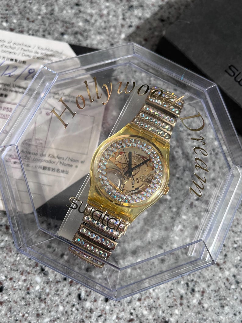 May include: A gold-tone Swatch watch with a clear face and a crystal-encrusted band. The watch is in a clear plastic box with the words "Hollywood Dream" printed on the side.