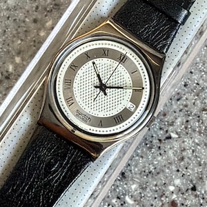 K&ouml;nnte beinhalten: Eine Swatch Swiss-Uhr mit einem schwarzen Lederarmband. Das Zifferblatt ist wei&szlig; mit einem strukturierten Zentrum und r&ouml;mischen Ziffern als Stundenmarkierungen. Die Uhr hat eine Datumsanzeige bei 3 Uhr und schwarze Zeiger.