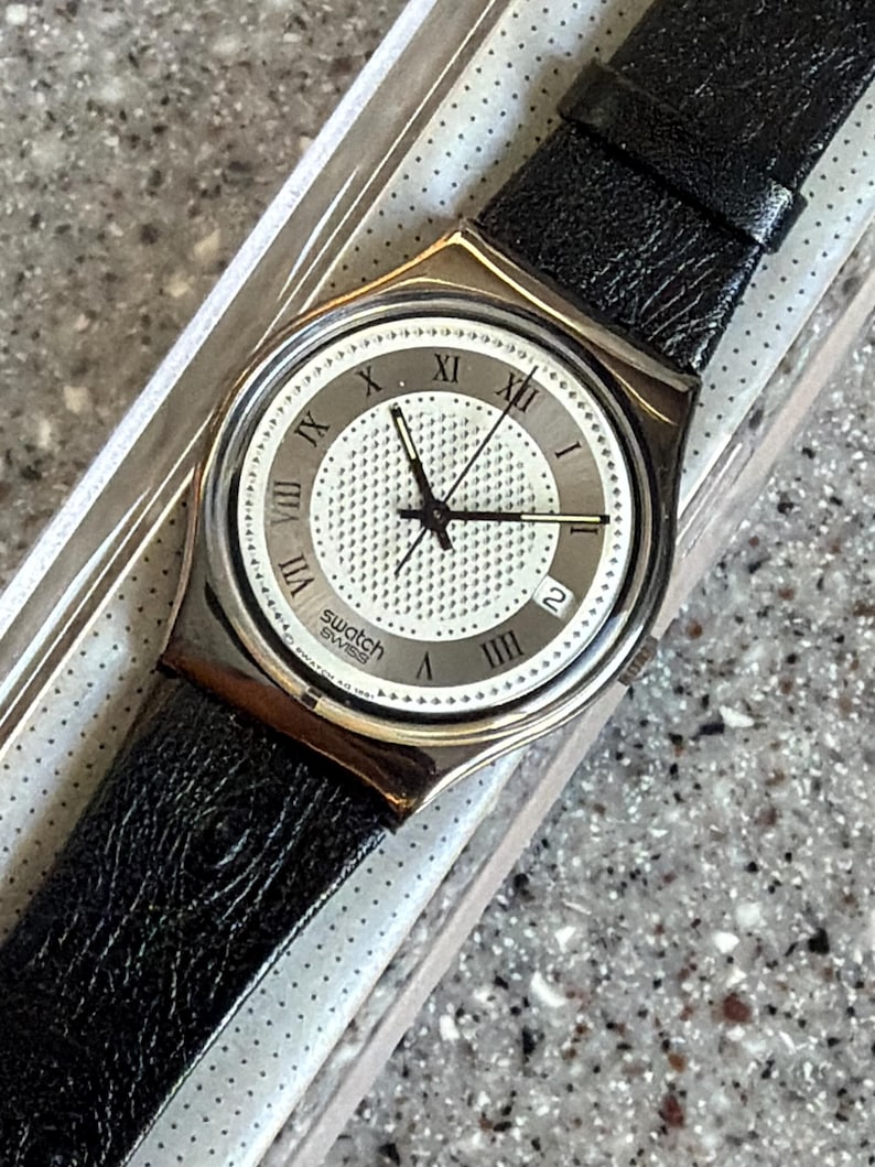 K&ouml;nnte beinhalten: Eine Swatch Swiss-Uhr mit einem schwarzen Lederarmband. Das Zifferblatt ist wei&szlig; mit einem strukturierten Zentrum und r&ouml;mischen Ziffern als Stundenmarkierungen. Die Uhr hat eine Datumsanzeige bei 3 Uhr und schwarze Zeiger.