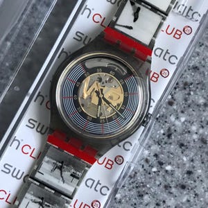 Op de afbeelding: Een zwart en zilveren Swatch-horloge met een transparante kunststof behuizing. De wijzerplaat van het horloge heeft een zwart-wit ontwerp met een gouden binnenring en een zichtbaar mechanisch uurwerk. Het horloge bevindt zich in een transparante kunststof display met een rode band en witte tekst die "Swatch Club 25" en "atc" luidt.