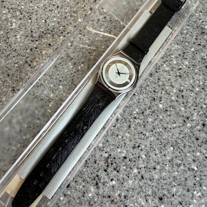 K&ouml;nnte beinhalten: Eine Swatch-Uhr mit schwarzem Lederarmband und silberfarbenem Geh&auml;use. Das Zifferblatt der Uhr ist wei&szlig; mit schwarzen Zeigern und r&ouml;mischen Ziffern. Die Uhr befindet sich in ihrer originalen transparenten Kunststoffverpackung.