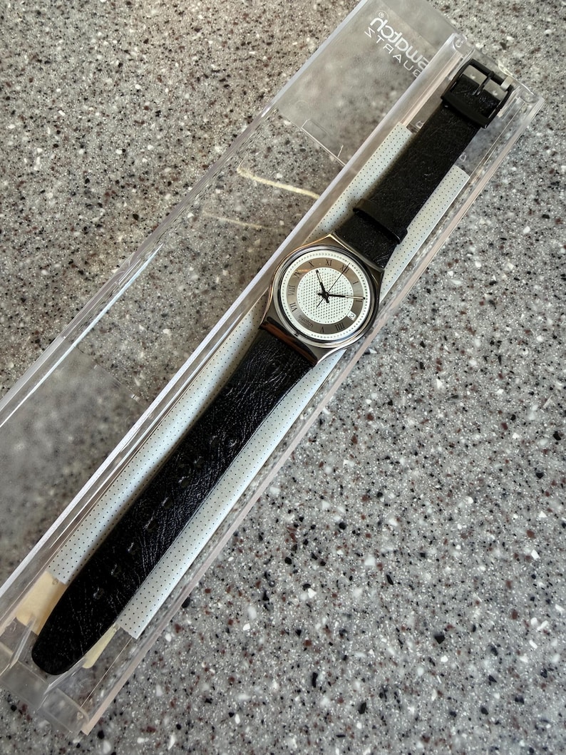 K&ouml;nnte beinhalten: Eine Swatch-Uhr mit schwarzem Lederarmband und silberfarbenem Geh&auml;use. Das Zifferblatt der Uhr ist wei&szlig; mit schwarzen Zeigern und r&ouml;mischen Ziffern. Die Uhr befindet sich in ihrer originalen transparenten Kunststoffverpackung.