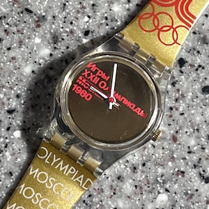 Puede incluir: Un reloj de pulsera de plástico transparente con una banda dorada. La banda tiene los anillos olímpicos y el texto "OLYMPIAD MOSCOW MOSCOW MOSKBA". La esfera del reloj es negra con manecillas blancas y el texto "Игры XXII Олимпиады 1980" en rojo.