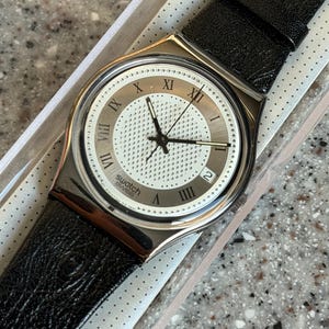 K&ouml;nnte beinhalten: Eine Swatch-Uhr mit silberfarbenem Geh&auml;use und wei&szlig;em Zifferblatt mit r&ouml;mischen Ziffern und einem strukturierten Zentrum. Die Uhr hat schwarze Zeiger und ein schwarzes Lederarmband. Die Uhr befindet sich in der Originalverpackung.