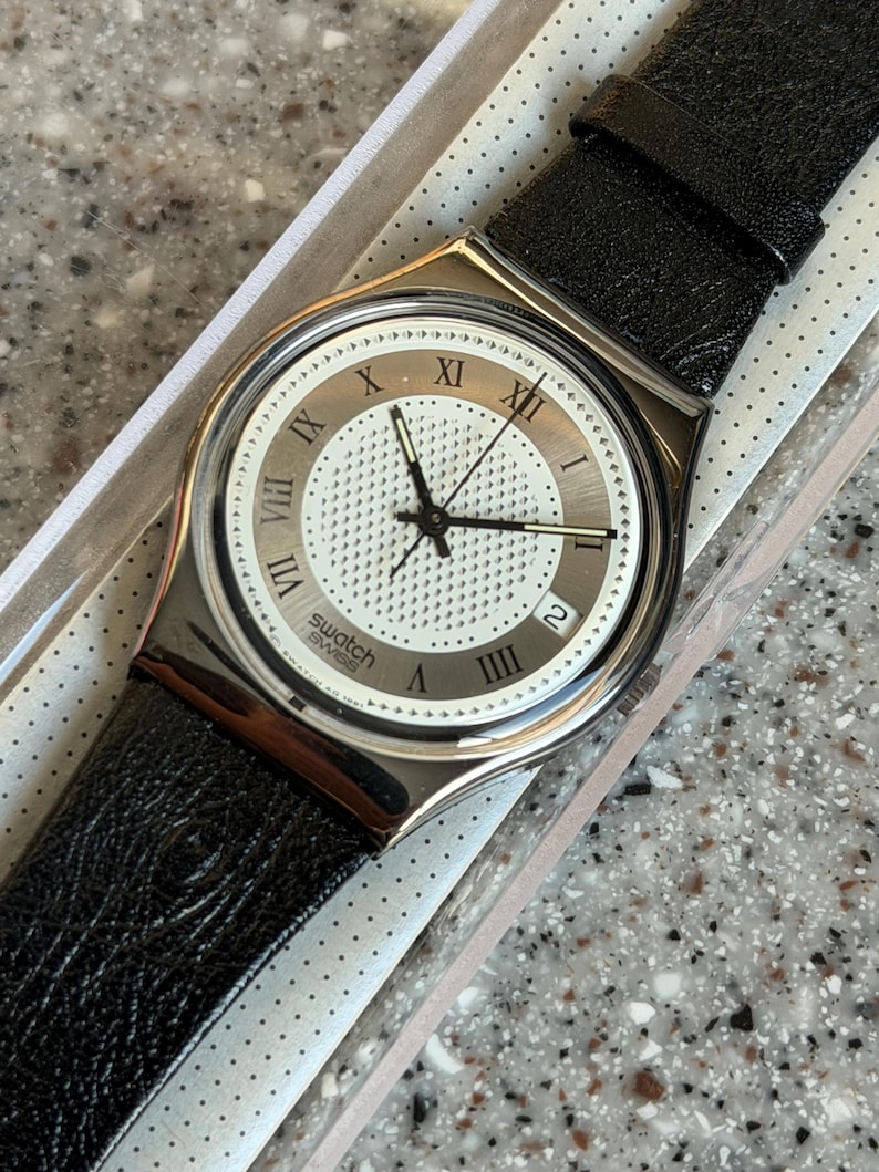 K&ouml;nnte beinhalten: Eine Swatch-Uhr mit silberfarbenem Geh&auml;use und wei&szlig;em Zifferblatt mit r&ouml;mischen Ziffern und einem strukturierten Zentrum. Die Uhr hat schwarze Zeiger und ein schwarzes Lederarmband. Die Uhr befindet sich in der Originalverpackung.