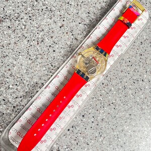 May include: A red wristwatch with a transparent face and a gold-colored buckle. The watch has a red strap and is displayed in its original packaging. The watch face shows the inner workings of the watch.