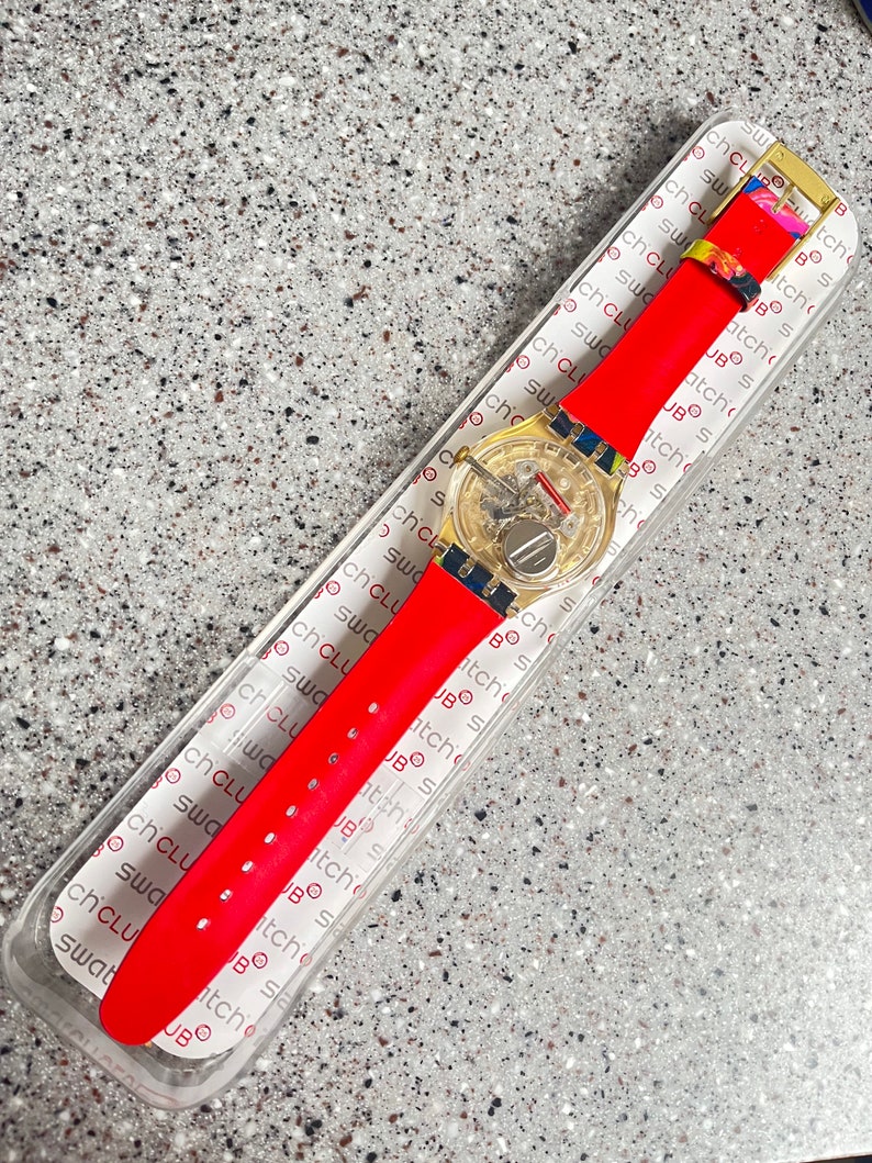 May include: A red wristwatch with a transparent face and a gold-colored buckle. The watch has a red strap and is displayed in its original packaging. The watch face shows the inner workings of the watch.