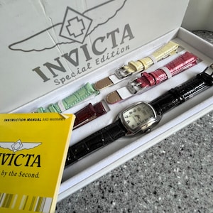 Puede incluir: Una caja blanca con el logotipo de Invicta Special Edition. La caja contiene cuatro correas de reloj de diferentes colores y un reloj plateado con una esfera blanca.