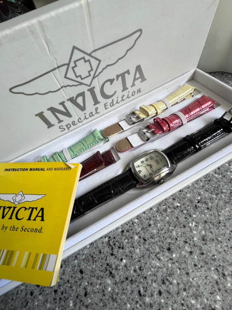 Puede incluir: Una caja blanca con el logotipo de Invicta Special Edition. La caja contiene cuatro correas de reloj de diferentes colores y un reloj plateado con una esfera blanca.