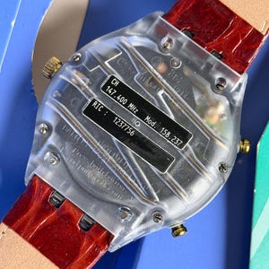 May include: A clear, rectangular watch with a red leather band. The watch face has a black label with text including "CH 147.400 MHz" and "Mod. 158.237". The watch has gold-colored accents.