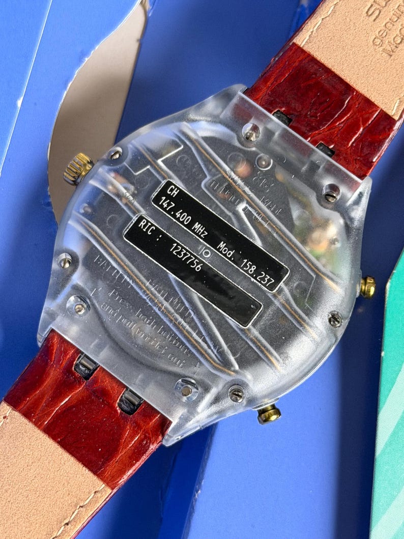 May include: A clear, rectangular watch with a red leather band. The watch face has a black label with text including "CH 147.400 MHz" and "Mod. 158.237". The watch has gold-colored accents.