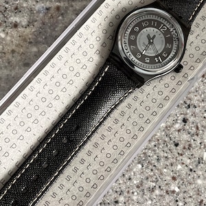 Puede incluir: Un reloj Swatch negro con una esfera blanca y agujas negras. El reloj tiene una correa de cuero negro con costuras blancas. La esfera del reloj tiene la palabra "Stop" impresa.