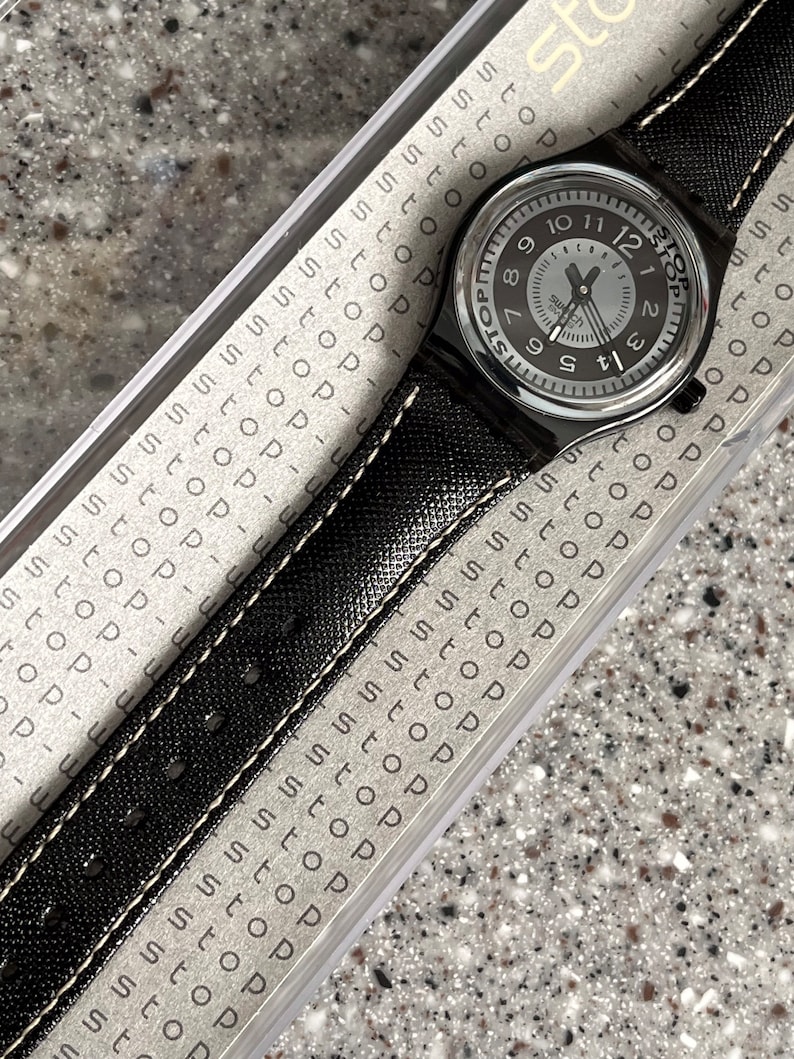 Puede incluir: Un reloj Swatch negro con una esfera blanca y agujas negras. El reloj tiene una correa de cuero negro con costuras blancas. La esfera del reloj tiene la palabra "Stop" impresa.