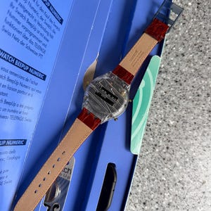 May include: A Swatch BeepUp Numeric watch with a clear case and a red leather strap. The watch is displayed in its original blue box, with text in multiple languages. The watch has a digital display.