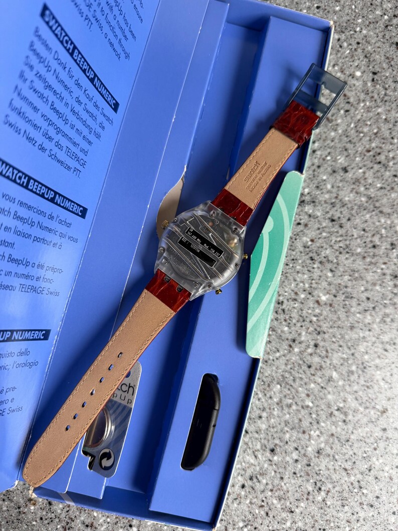 May include: A Swatch BeepUp Numeric watch with a clear case and a red leather strap. The watch is displayed in its original blue box, with text in multiple languages. The watch has a digital display.