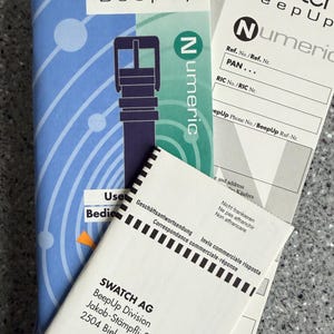 May include: A stack of Swatch BeepUp watch instruction manuals and paperwork. The main booklet is blue with a watch strap illustration. Additional paperwork includes the "Numeric" logo and address information for SWATCH AG.