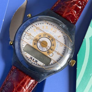 May include: A Swatch watch with a white face, gold accents, and a digital display. The watch has a black case and a red leather band. The watch face displays the text "Swatch Swiss". The watch is in its original packaging.
