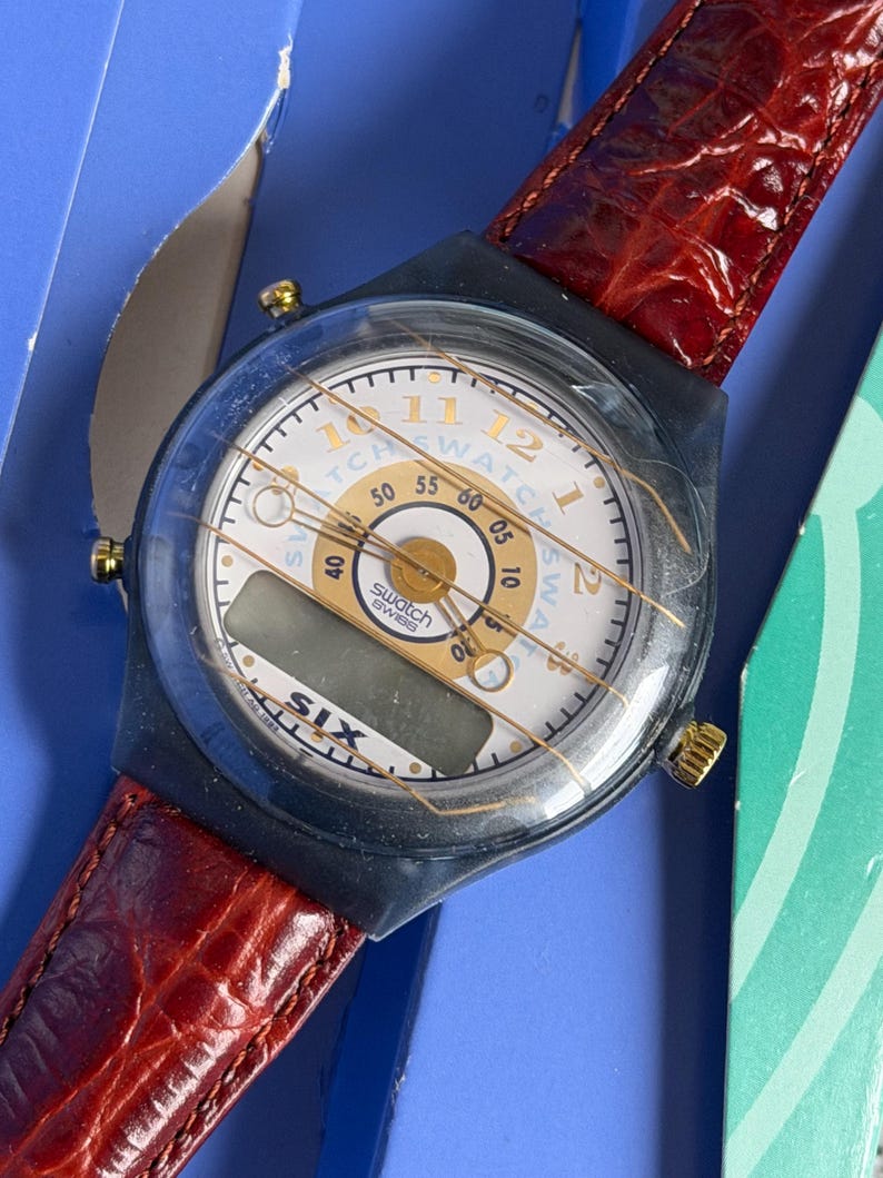 May include: A Swatch watch with a white face, gold accents, and a digital display. The watch has a black case and a red leather band. The watch face displays the text "Swatch Swiss". The watch is in its original packaging.