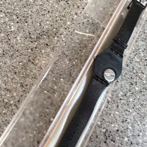 K&ouml;nnte beinhalten: Eine schwarze Armbanduhr mit Lederarmband wird in einer transparenten Kunststoffbox pr&auml;sentiert. Die Uhr hat ein rundes schwarzes Zifferblatt mit silbernen Akzenten und eine Schnalle. Auf dem Geh&auml;use steht der Text "notowe STRAUE".
