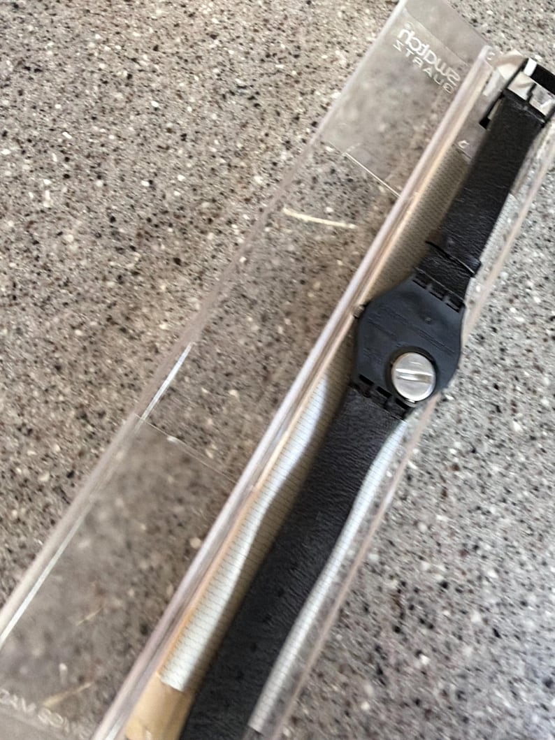 K&ouml;nnte beinhalten: Eine schwarze Armbanduhr mit Lederarmband wird in einer transparenten Kunststoffbox pr&auml;sentiert. Die Uhr hat ein rundes schwarzes Zifferblatt mit silbernen Akzenten und eine Schnalle. Auf dem Geh&auml;use steht der Text "notowe STRAUE".