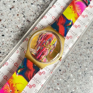 May include: A gold-toned Swatch watch with a colorful abstract design on the face and strap. The watch face features a portrait with vibrant hues of red, yellow, and blue. The strap has a matching pattern. The watch is in its original packaging.