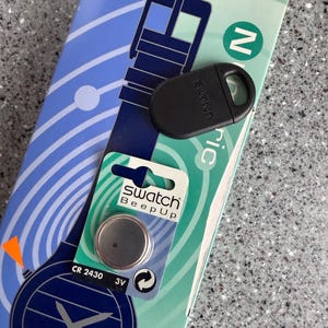 May include: A Swatch Beep Up watch battery and black watch accessory are displayed on a patterned blue and green background. The battery is a CR 2430 3V. The watch accessory is black and has the Swatch logo.
