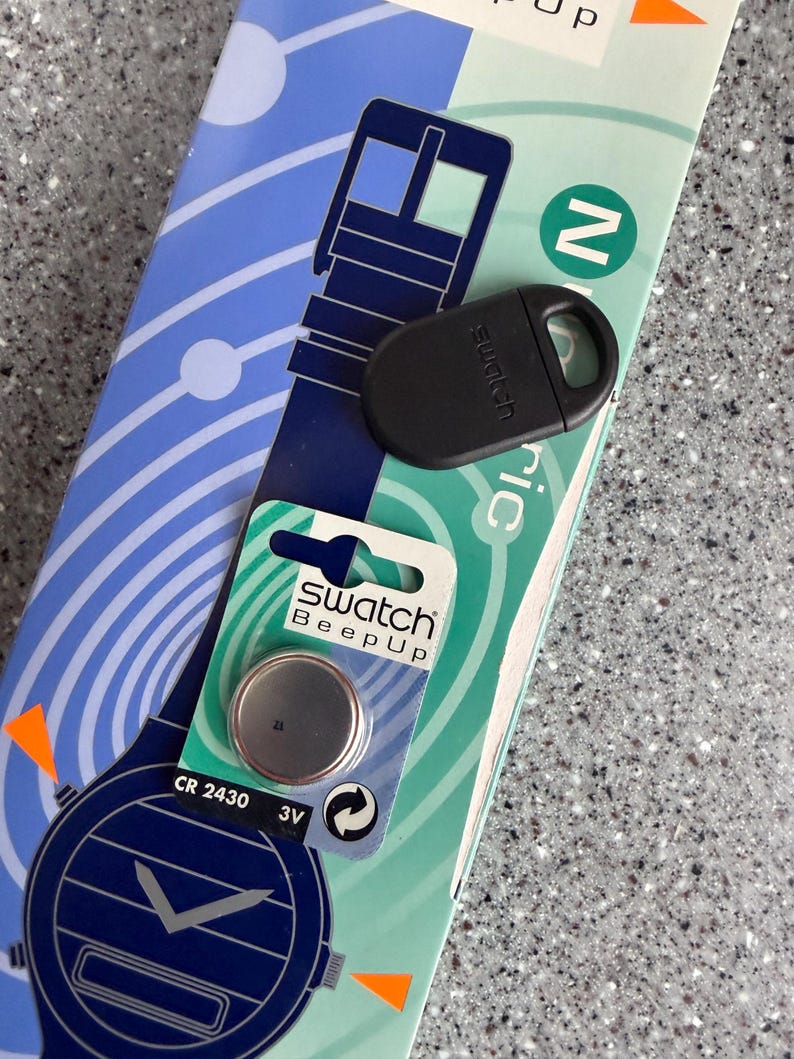 May include: A Swatch Beep Up watch battery and black watch accessory are displayed on a patterned blue and green background. The battery is a CR 2430 3V. The watch accessory is black and has the Swatch logo.