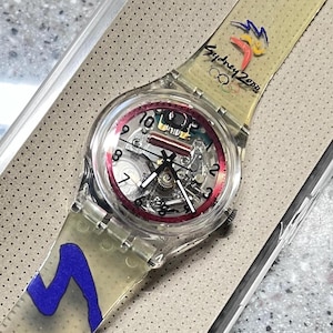 Könnte beinhalten: Eine transparente Armbanduhr aus Kunststoff mit einem roten und schwarzen Zifferblatt. Das Armband der Uhr hat ein blaues und gelbes Design und den Text "Sydney 2000" mit den olympischen Ringen.