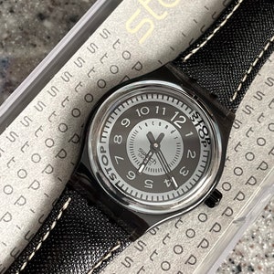 Puede incluir: Un reloj Swatch negro con una esfera blanca y agujas negras. El reloj tiene una correa de cuero negro con costuras blancas. El reloj est&aacute; en una caja de pl&aacute;stico transparente con la palabra "Stop" impresa en ella.