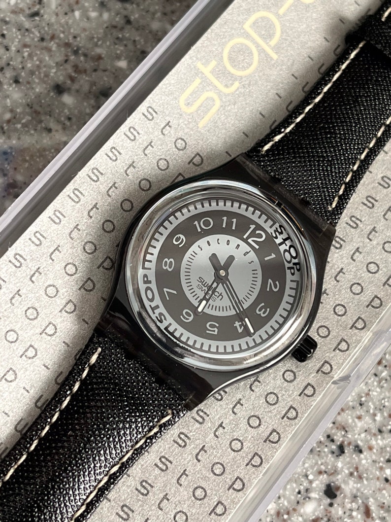 Puede incluir: Un reloj Swatch negro con una esfera blanca y agujas negras. El reloj tiene una correa de cuero negro con costuras blancas. El reloj est&aacute; en una caja de pl&aacute;stico transparente con la palabra "Stop" impresa en ella.