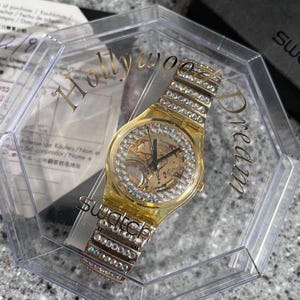 May include: A gold-tone Swatch watch with a clear crystal face and a crystal-encrusted band. The watch is in a clear plastic box with the words "Hollywood Dream" printed on the side.