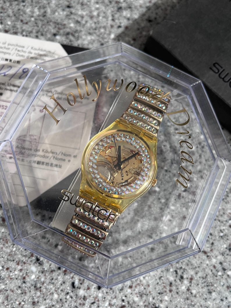 May include: A gold-tone Swatch watch with a clear crystal face and a crystal-encrusted band. The watch is in a clear plastic box with the words "Hollywood Dream" printed on the side.