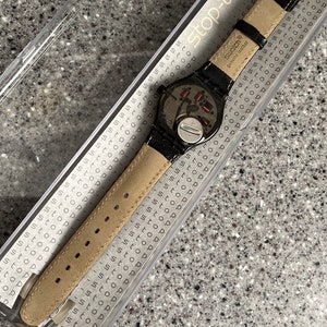 Puede incluir: Un reloj Swatch negro con una correa de cuero negro y una esfera plateada. El reloj est&aacute; en una caja de pl&aacute;stico transparente con las palabras "stop-watch" impresas en el lateral. La correa del reloj tiene las palabras "swatch&reg; genuine leather" impresas en ella.