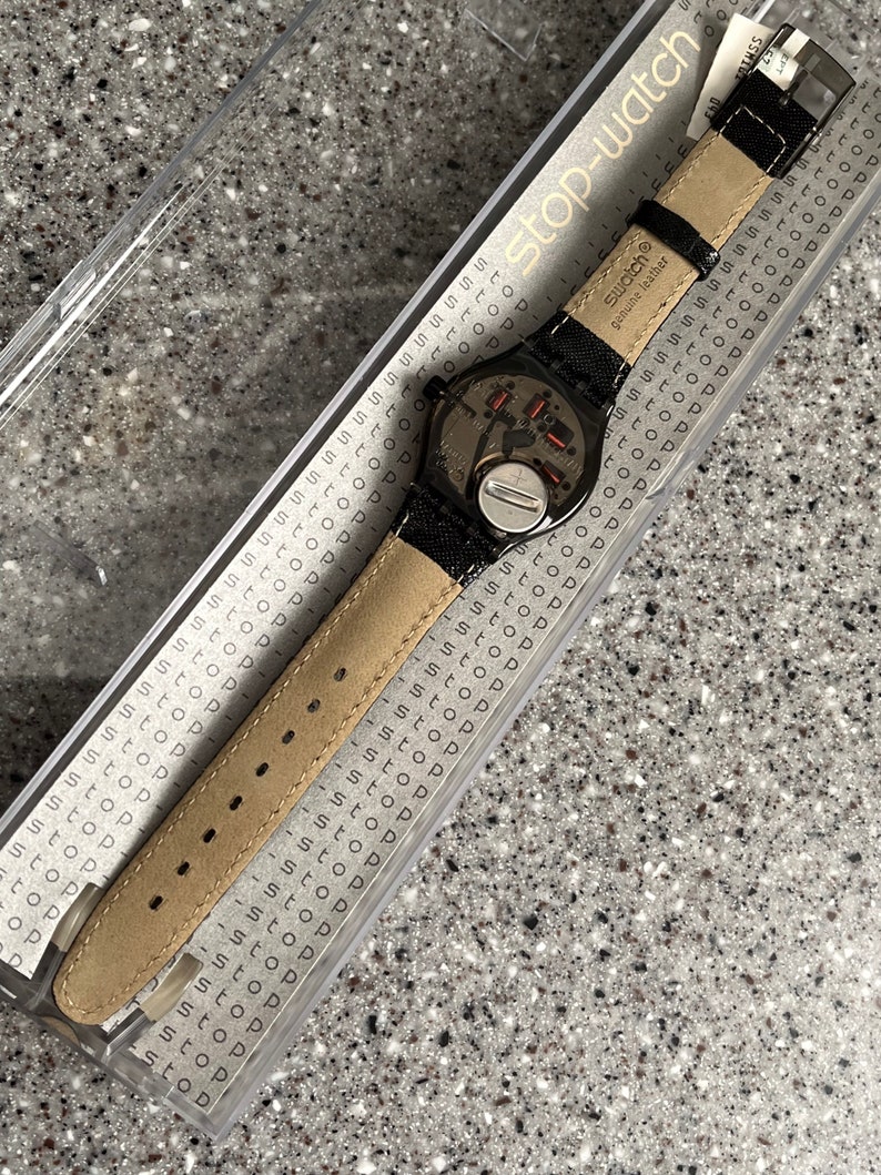 Puede incluir: Un reloj Swatch negro con una correa de cuero negro y una esfera plateada. El reloj est&aacute; en una caja de pl&aacute;stico transparente con las palabras "stop-watch" impresas en el lateral. La correa del reloj tiene las palabras "swatch&reg; genuine leather" impresas en ella.