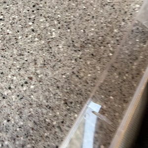 K&ouml;nnte beinhalten: Nahaufnahme einer gesprenkelten grauen Oberfl&auml;che mit einem transparenten Kunststoffbeh&auml;lter in der Ecke. Die Oberfl&auml;che hat ein Muster aus kleinen schwarzen, wei&szlig;en und grauen Punkten. Der Beh&auml;lter ist teilweise sichtbar, mit einem wei&szlig;en Etikett.