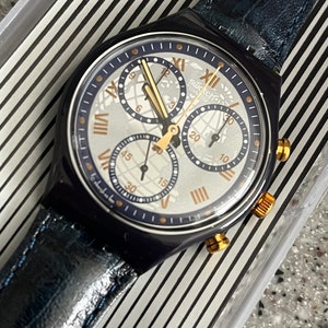 May include: A blue and gold Swatch chronograph watch with a white face and a blue leather band. The watch has a world map design and Roman numeral hour markers.