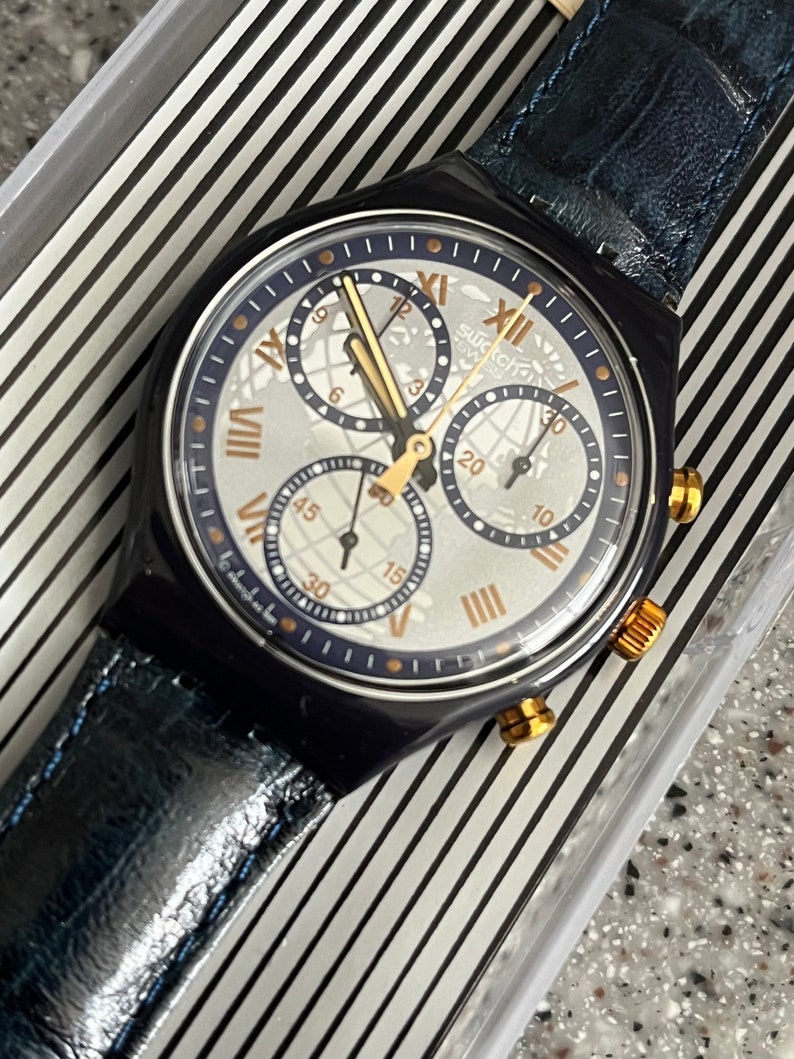 May include: A blue and gold Swatch chronograph watch with a white face and a blue leather band. The watch has a world map design and Roman numeral hour markers.