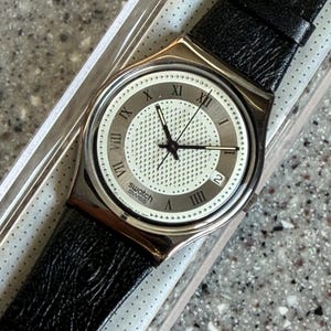 K&ouml;nnte beinhalten: Eine Swatch Swiss Uhr mit silberfarbenem Geh&auml;use und schwarzem Lederarmband. Das Zifferblatt ist wei&szlig; mit silbernen r&ouml;mischen Ziffern und einem Datumsfenster. Die Uhr befindet sich in der Originalverpackung.