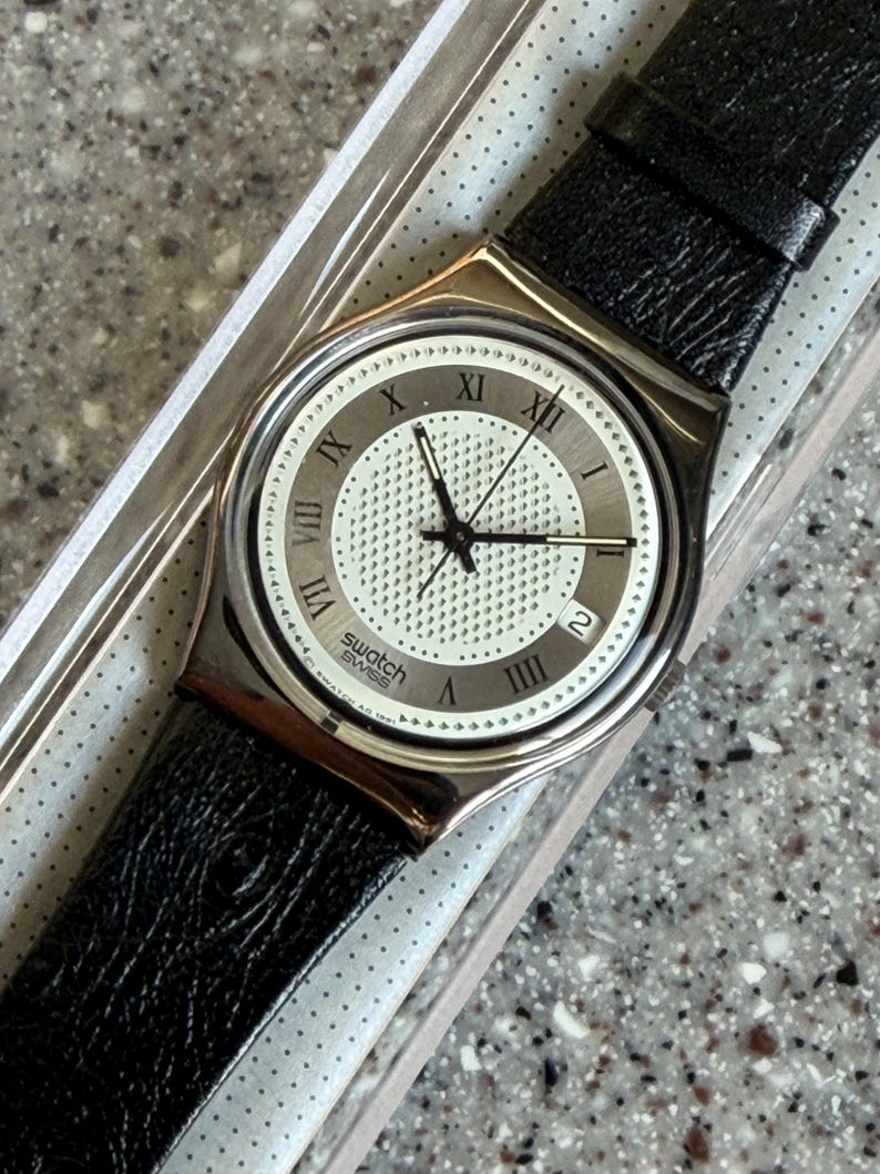 K&ouml;nnte beinhalten: Eine Swatch Swiss Uhr mit silberfarbenem Geh&auml;use und schwarzem Lederarmband. Das Zifferblatt ist wei&szlig; mit silbernen r&ouml;mischen Ziffern und einem Datumsfenster. Die Uhr befindet sich in der Originalverpackung.