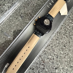 May include: A black Swatch watch with a black leather strap and gold accents. The watch face is black with white numbers and hands. The watch is in a clear plastic box with a black and white striped background. The box has the word "CHRONO" printed on it.