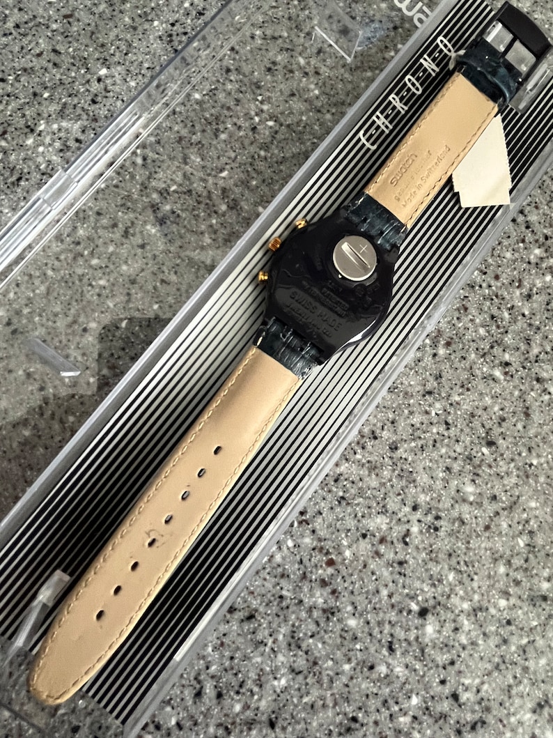 May include: A black Swatch watch with a black leather strap and gold accents. The watch face is black with white numbers and hands. The watch is in a clear plastic box with a black and white striped background. The box has the word "CHRONO" printed on it.
