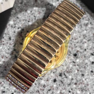 May include: Gold-tone watch with a gold-tone expandable band with clear crystals. The band has a ribbed texture.
